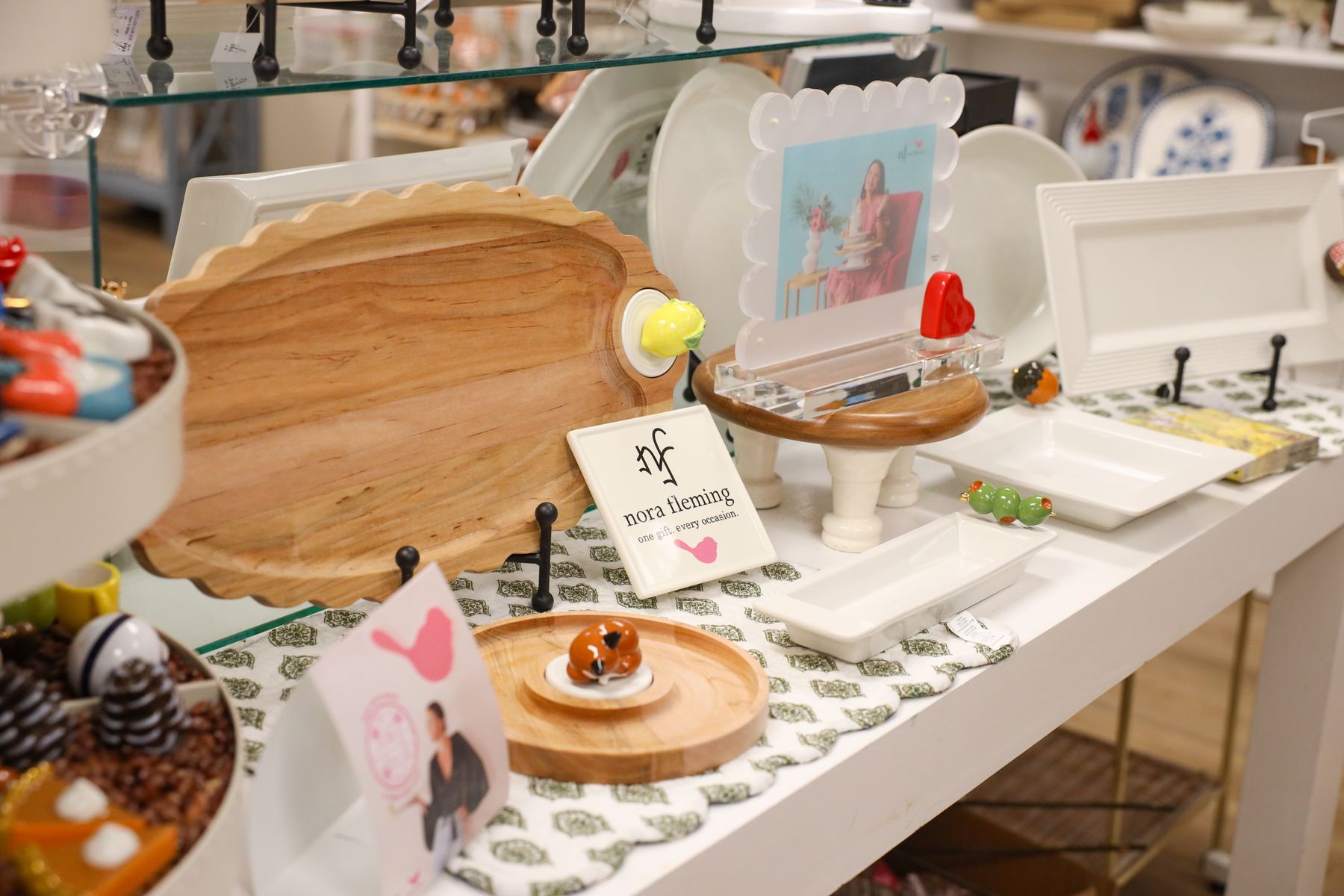 A wooden serving board, Nora Fleming serving pieces, and small dishes on a white display table