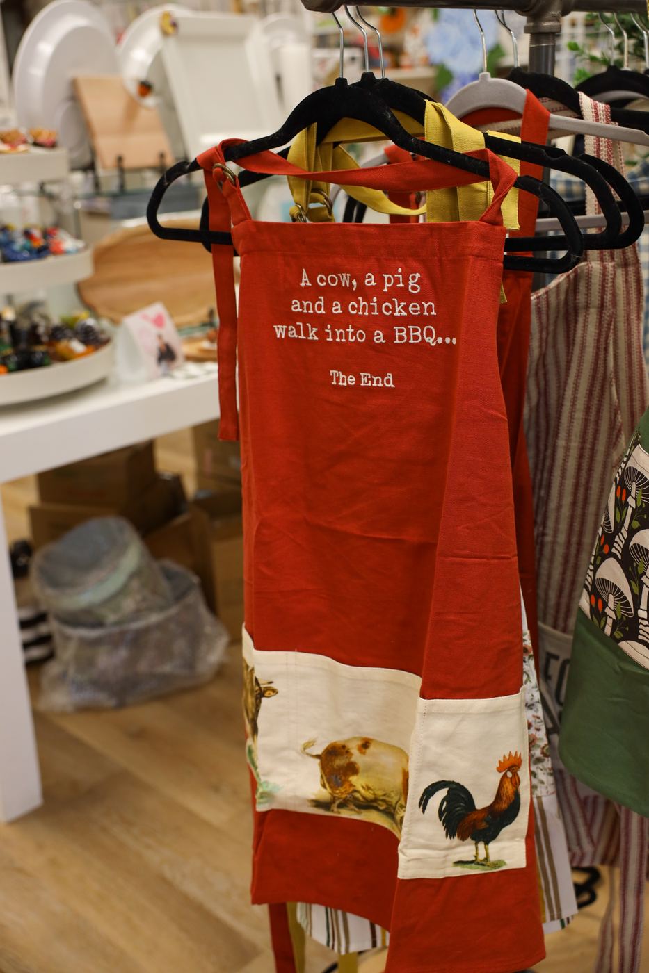 A red apron printed with a farm joke and a rooster, hanging with other aprons
