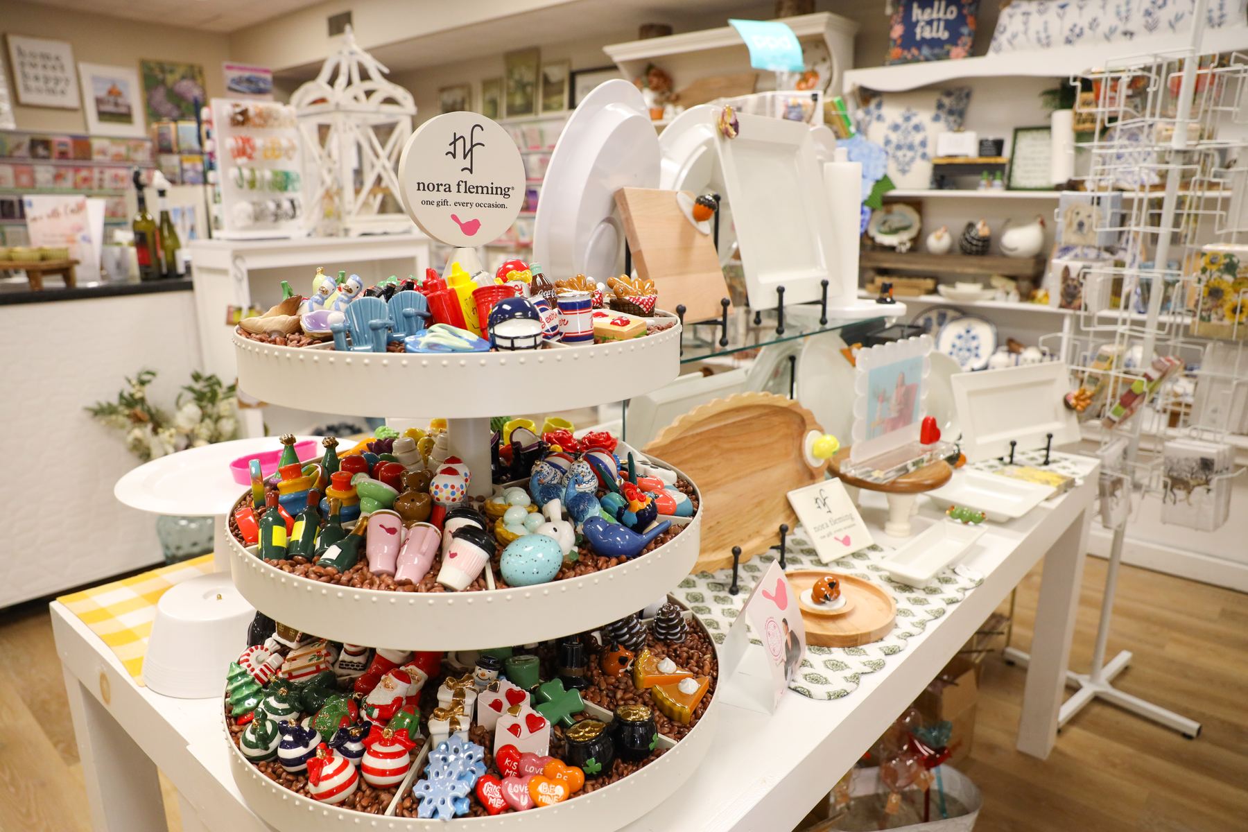 A tiered Nora Fleming display filled with minis beside white serving platters