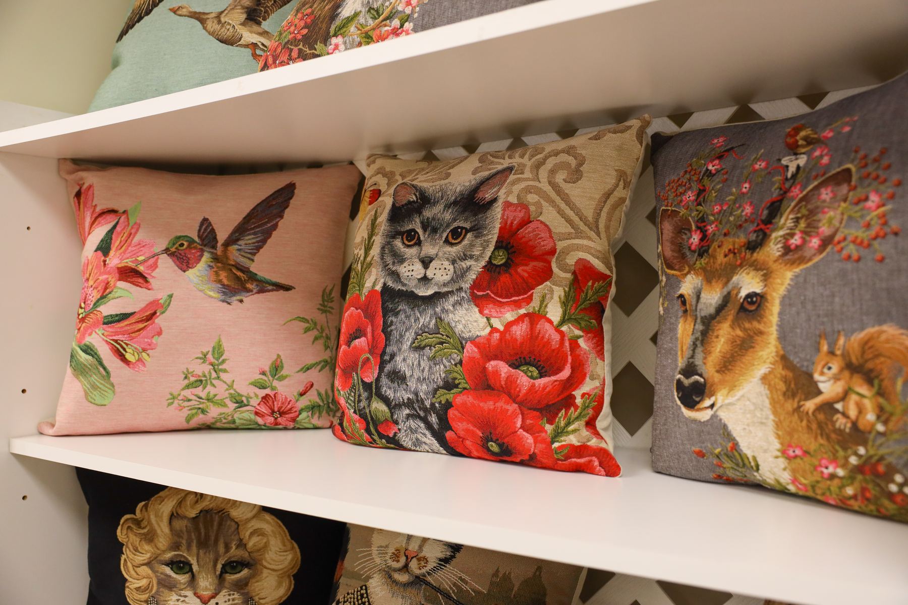Tapestry pillows of a hummingbird, a gray cat among red poppies, and a deer on a white shelf