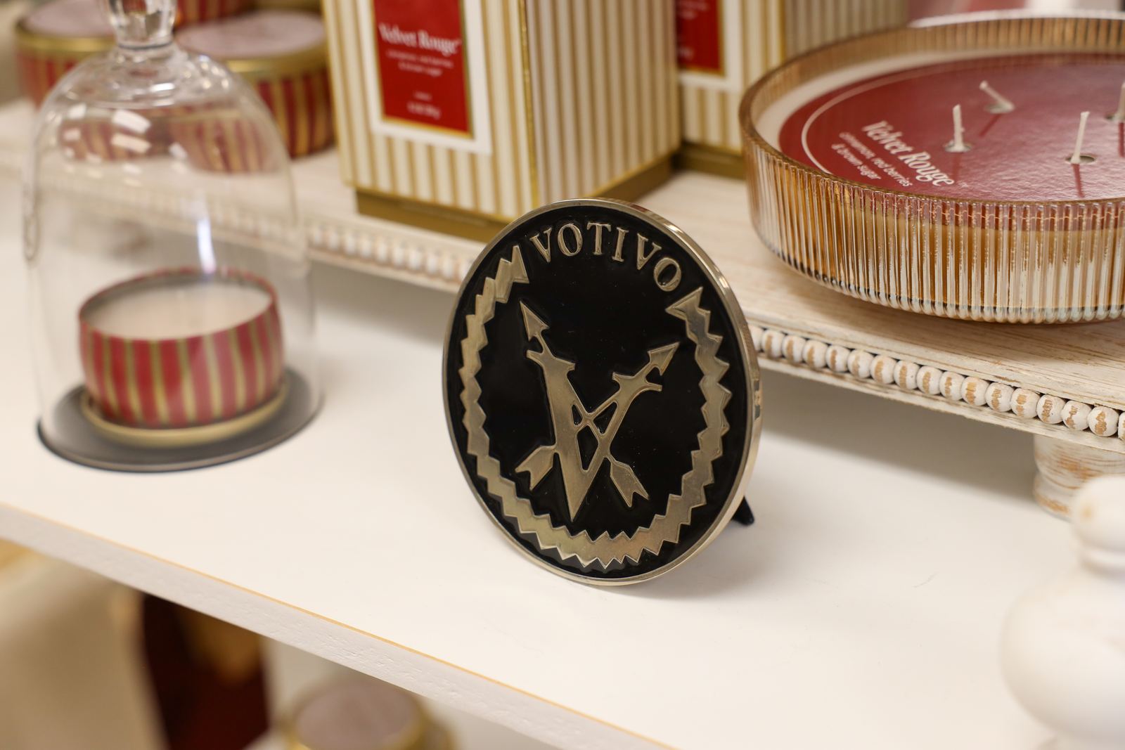 Votivo Velvet Rouge candles on a shop shelf, one under a glass cloche, beside the Votivo medallion
